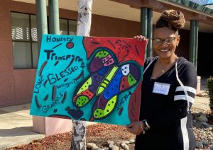 Women's Empowerment Graduate in WE's front yard, showing off her painting of a butterfly, with descriptive and motivational words on the canvas around the butterfly "honesty, transformation, blessed, grateful, love"