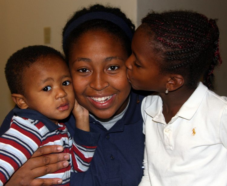 Three young children, a young boy staring at the camera on the left, a young girl in the middle smiling at the camera, and a young girl on the right kissing the cheek of the middle girl.