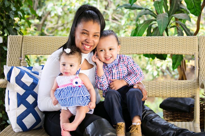 Women's Empowerment graduate, Natoshi, posing for the camera on a chair, holding her two kids, dressed in matching outfits. All smiling at the camera.